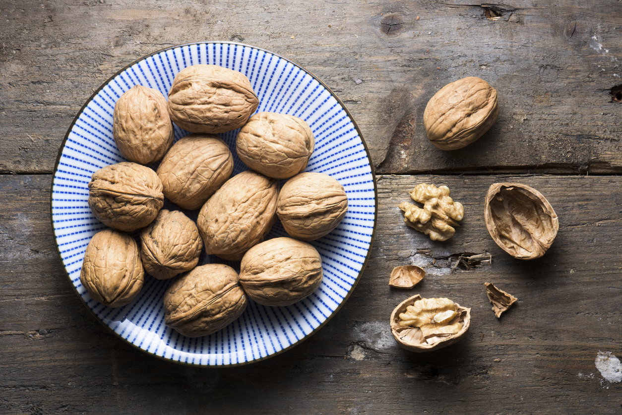 A bowl of walnuts is sitting on a rough wooden table. There are loose walnuts beside the bowl. One nut is cracked open, surrounded by the parts of its shell. The bowl is blue and white striped and casts a strong shadow onto the textured surface beneath it.